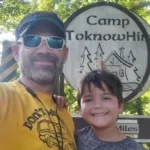 Jay and his son standing together, smiling, in front of the Camp Toknowhim sign, capturing a joyful moment and their connection to the camp.