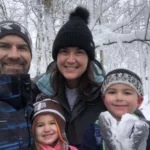 Jay Dryden and his family smiling happily in an outdoor wood area covered in snow, enjoying the winter scenery and creating joyful memories together.
