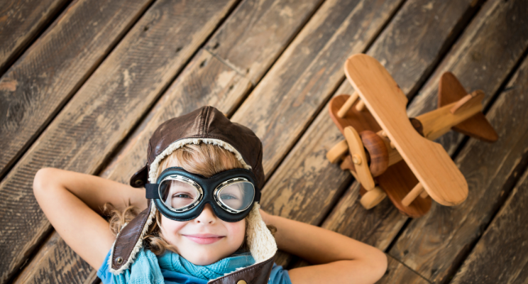 A child wearing airplane goggles standing next to a wooden airplane model, evoking a sense of adventure, imagination, and fascination with aviation.