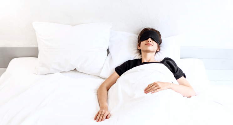 A woman lying in bed wearing a black eye mask, indicating relaxation, restfulness, and an effort to block out light for a better sleep experience.