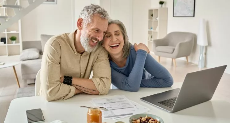 A happy couple with white and grey hair, smiling and seated in a modern living room, looking at a laptop screen. The image represents the concept of boosting retirement savings and planning for the future.
