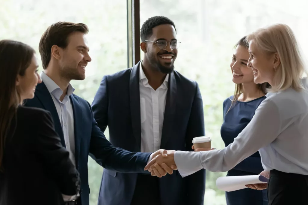 A diverse group of business professionals engaged in a lively conversation and exchanging handshakes in an office building.